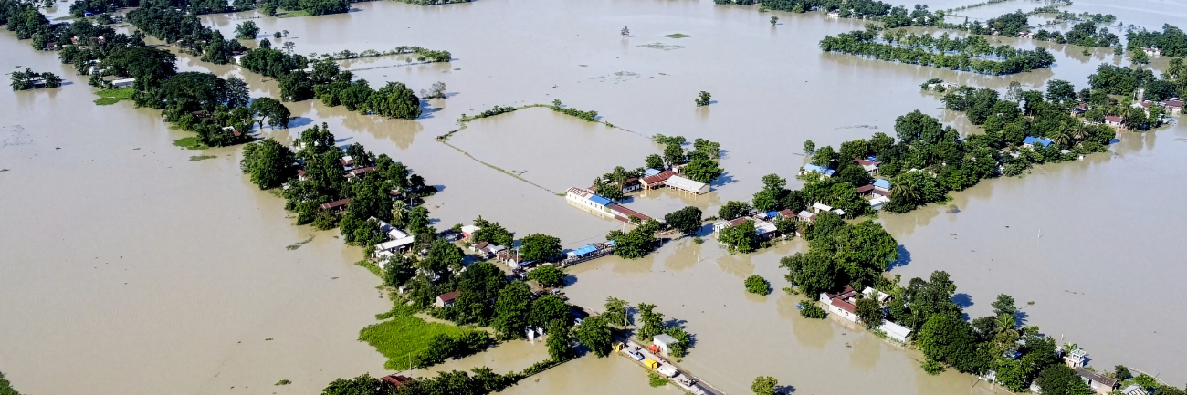 An aerial view shows widespread flooding submerging villages and farmland, with isolated homes and trees standing amid vast floodwaters