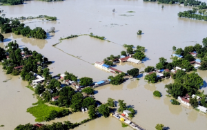 An aerial view shows widespread flooding submerging villages and farmland, with isolated homes and trees standing amid vast floodwaters