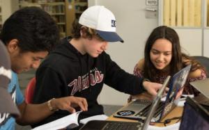 Three students gathering around a laptop and book