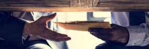 A picture of one hand passing an envelope to another hand under a table.